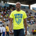 Man wearing a bright yellow 'Edwin Allen' t-shirt in an outdoor stadium setting. EAHS Men's Athletics Performance Tee – Front Logo & Back