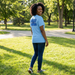 Woman wearing a light blue t-shirt with text in a park EAHS Edwin Allen Blue Tee - Crest Front - Built on Speed Back - Unisex T-Shirt